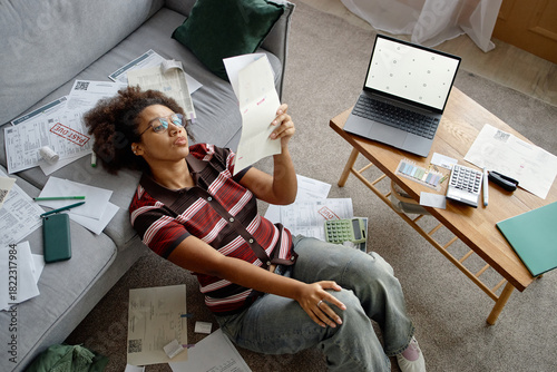 Young Black woman sitting on floor analyzing invoices and bills surrounded by paperwork using laptop and calculator financial documents scattered on table and couch
