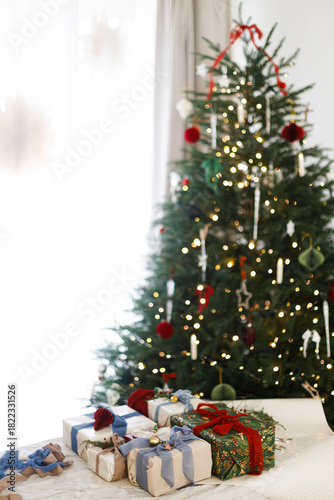  Merry Christmas and Happy Holidays! Wrapped stylish christmas presents with silk bows, green branch and berries, vintage ornaments on rustic table against decorated xmas tree with lights.