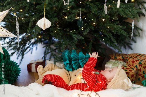 Merry Christmas and Happy Holidays! Cute baby in christmas costume playing with tree paper decoration and lying under stylish decorated christmas tree with gifts. Baby's first christmas