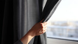 Close-up of a hand pulling back a gray curtain by a window with blurred outdoor background in soft natural light