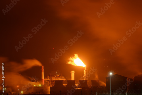 An industrial flare stack burns off excess gas with a large, bright orange flame and thick smoke