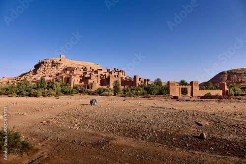 A view of Ait ben haddou, an ancient fortified village in Morocco. Tourists walk across the desert landscape to the Ksar on a clear, bright day.