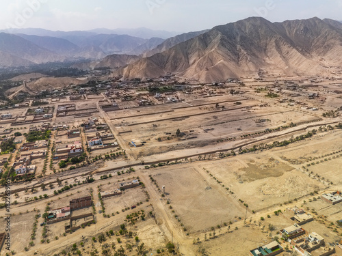 Aerial panoramic view of urban sprawl in the arid Cieneguilla valley near Lima Peru showing subdivided land plots and a dry riverbed against mountains.