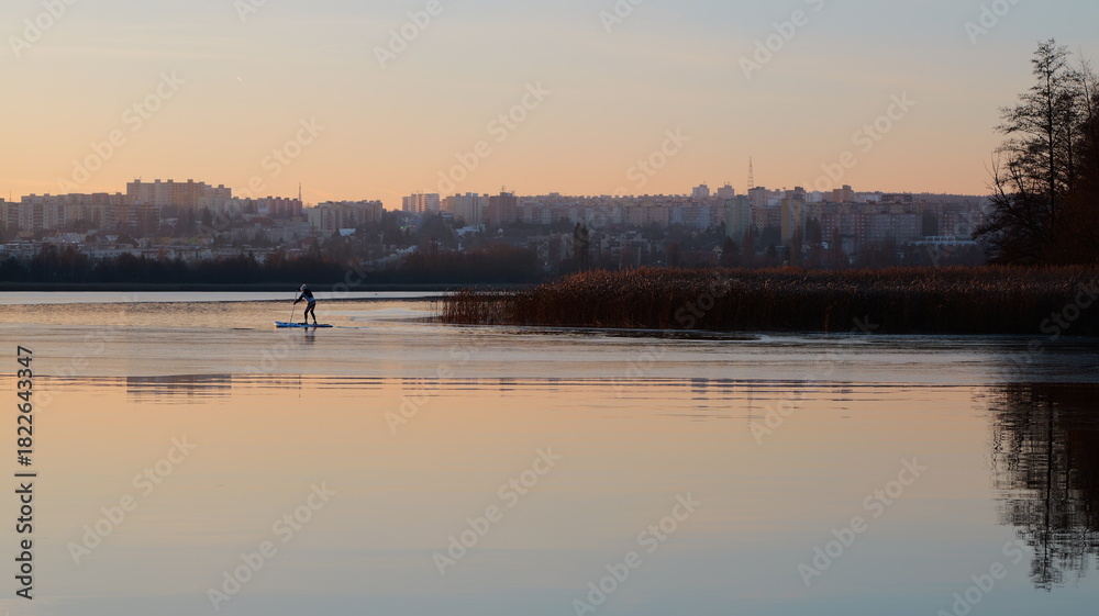 Naklejka premium A person paddles a board on water. A cityscape sits distant. Reeds line the shore. The sky glows orange. Reflections appear on the surface. Trees are visible. It is a ser