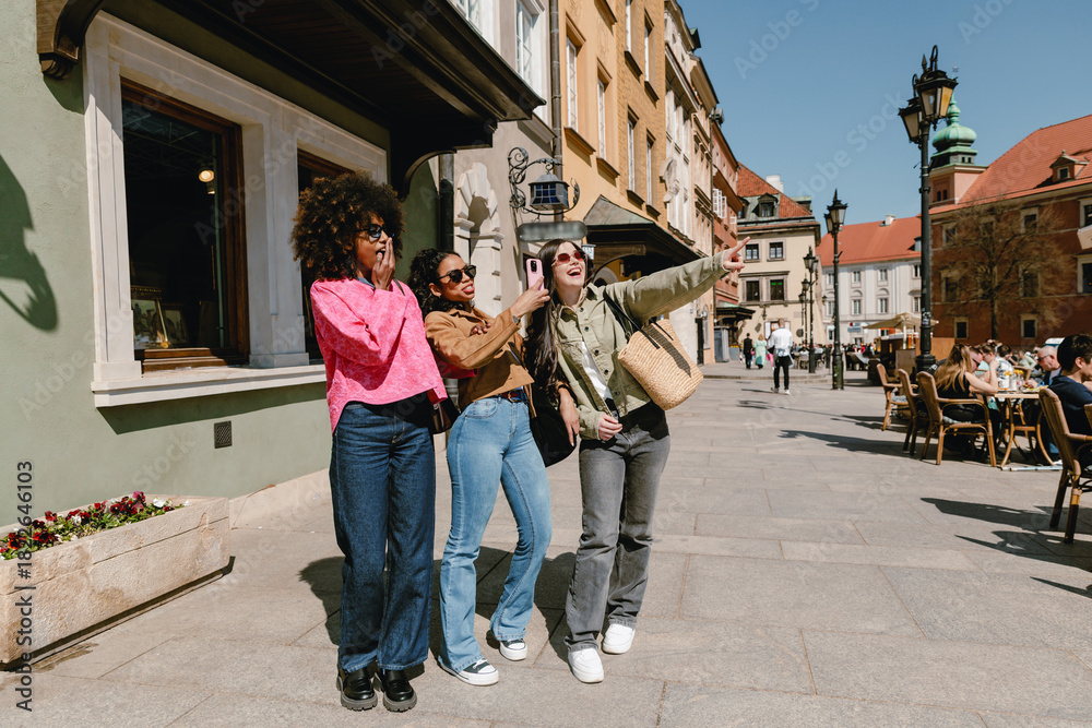 Fototapeta premium A group of three female friends stand and look in the direction one of them is pointing while one of them covers her mouth with her hand and the other holds a phone while they talk and laugh