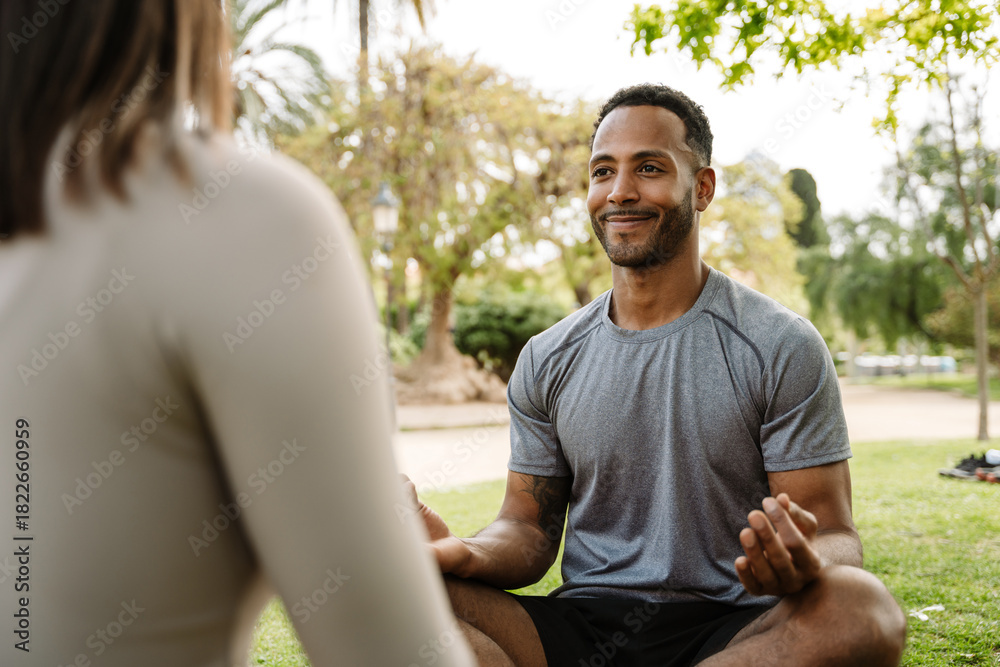 Fototapeta premium A male athlete is smiling and sitting on the grass with his legs crossed while looking at a female athlete across from him