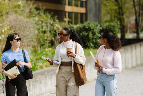 A female student holds a cup and a notebook while talking and listening to two female students walking next to her and holding folders