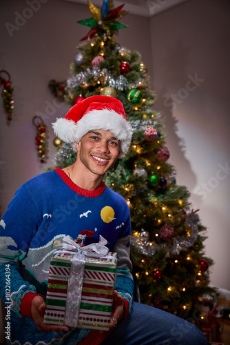 Man wearing Santa hat and festive sweater holding wrapped Christmas gift by decorated tree