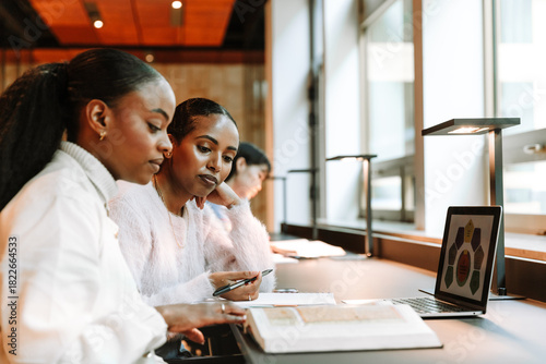 Two female students are reading a book while one of them holds a pen and they are sitting at a table in front of a laptop