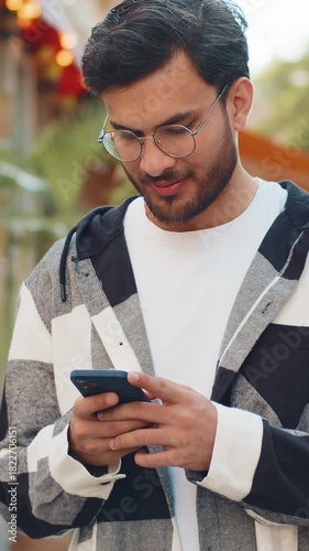 Indian young man using smartphone typing text messages in social media application online, surfing internet, relaxing, taking a break outdoors. Hispanic guy tourist standing on urban city street.
