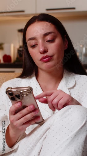 Young woman scrolls news at night home bored tired looks at smartphone sighs stressed unmotivated. Caucasian girl on sofa at evening shows fatigue burnout mood dull scroll stare poor focus wants rest.