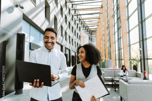 Male and female workers walking and laughing while he holds a laptop and she holds folders
