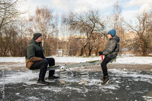 Adult and child sit on a seesaw in a snowy urban park, wearing winter coats and hats.