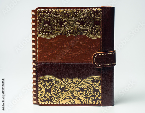 Leather journal with ornate gold detailing rests on a white surface, showcasing craftsmanship and vintage aesthetic, evoking a sense of timeless elegance.