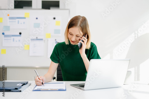 Business woman talking on the phone while writing notes and working on a laptop in a bright modern office.