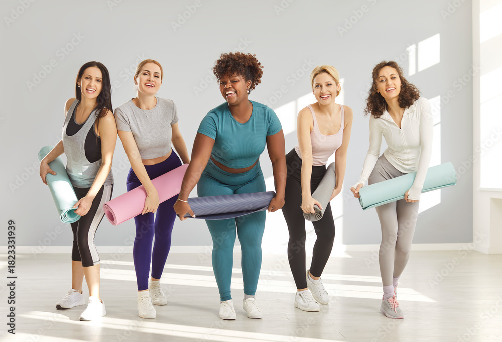 Fototapeta premium Group of multiracial happy women in sportswear posing together in bright fitness studio, holding yoga mats and smiling ready for yoga class. Active healthy lifestyle, friendship and wellness concept.