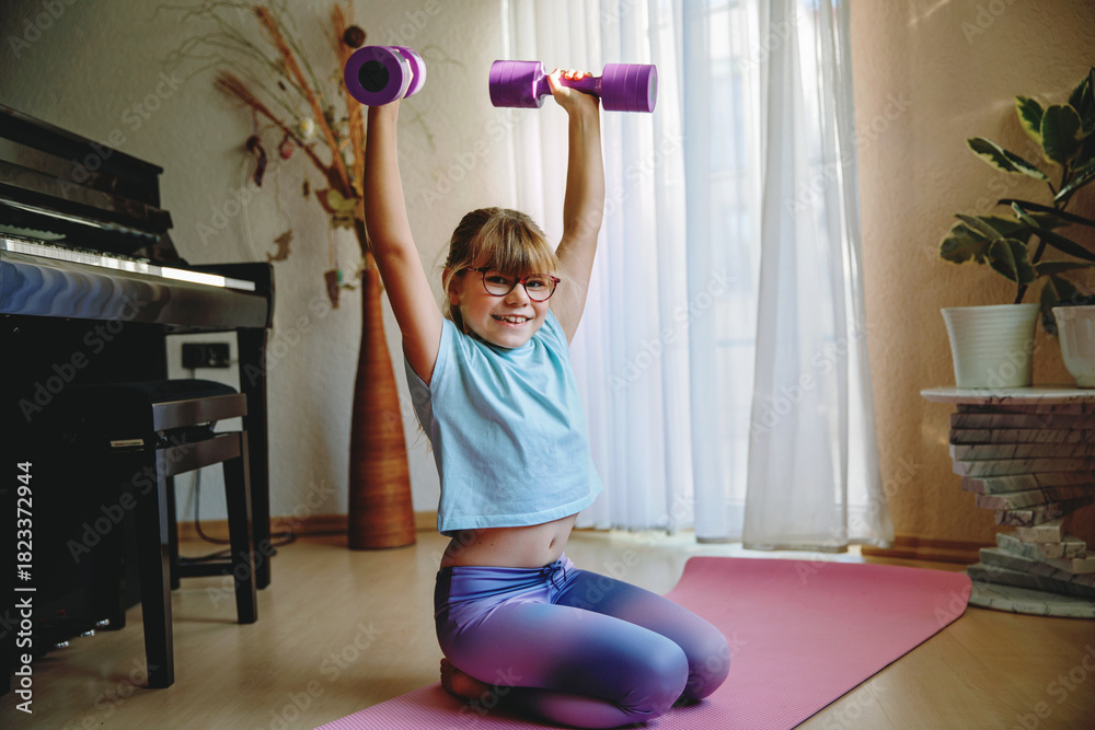 Fototapeta premium little girl trains at home with dumbbells, strength training and sports. Child having fun with training