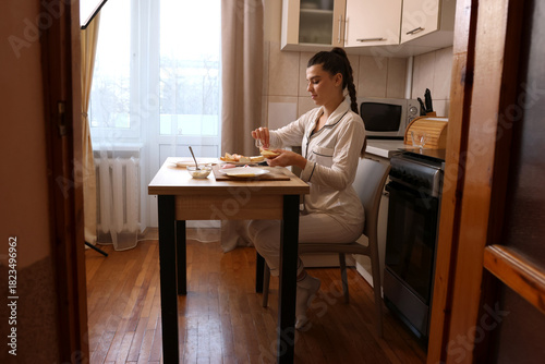 Beautiful young woman preparing sandwiches while sitting at the table in the kitchen.