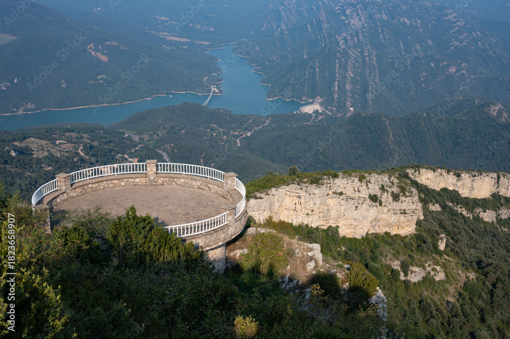 Obraz premium Circular viewing platform (mirador) with white railing, built high on a cliff, offering a stunning panoramic view of a reservoir and forested mountains near Berga, Catalonia