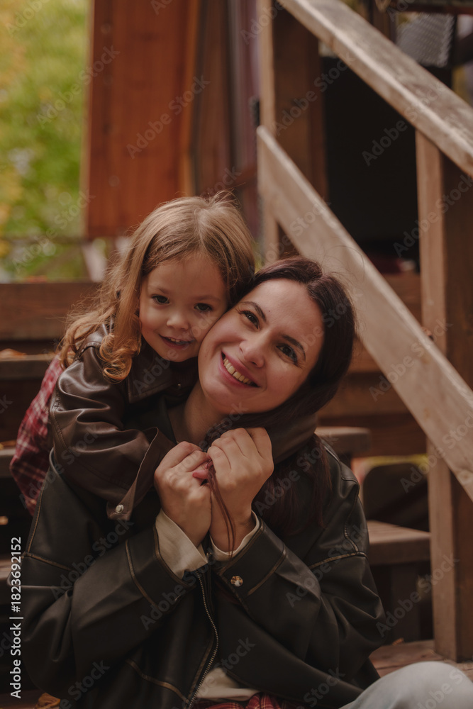 Fototapeta premium Mother and daughter hugging on the steps of a wooden staircase. Cheerful girls in the country in the fall. A little girl hugs her mother from behind. Rainy weather. Vertical photo.