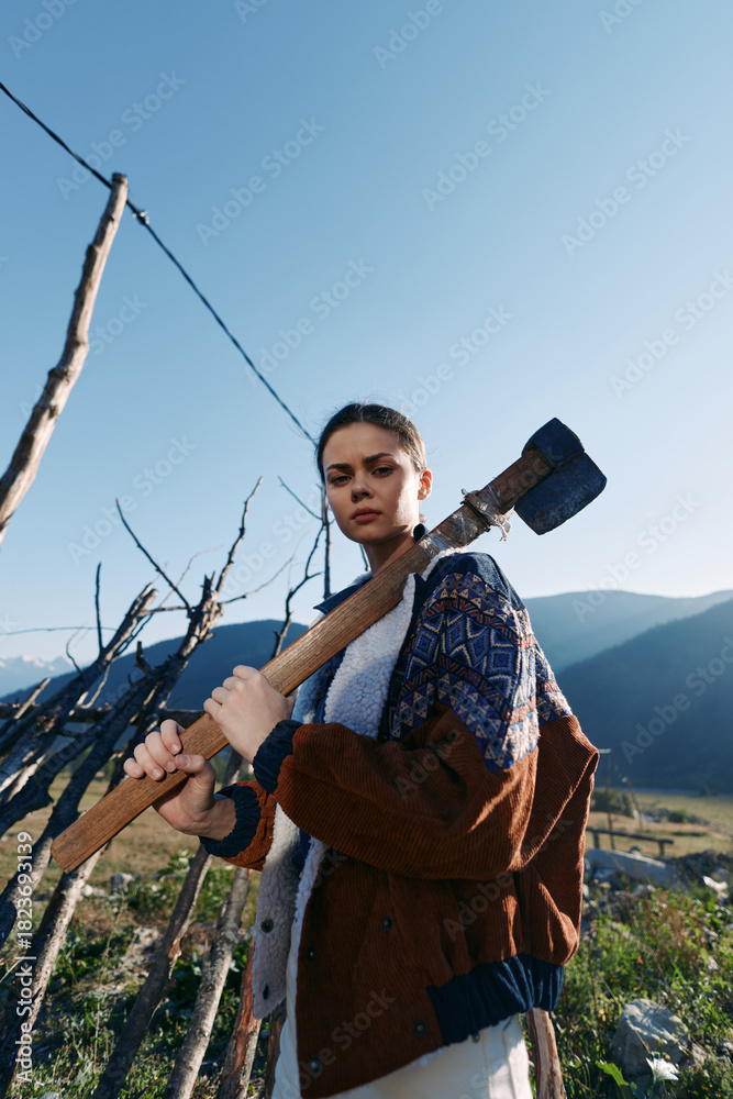 Naklejka premium Woman with axe outdoors in mountains rural fence tool nature, holding wooden handle by vineyard posts. Portrait of a young serious worker in rustic landscape with sunlight and determination.