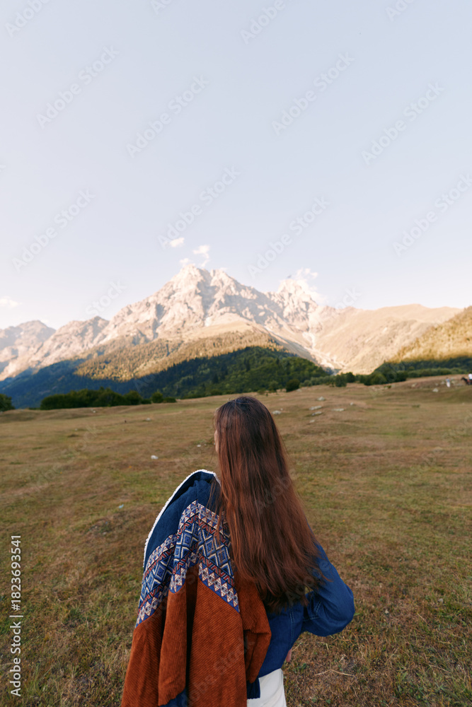 Fototapeta premium Woman with long hair from back wearing patterned shawl standing in meadow facing mountain landscape. Nature scene with wide grass field, outdoor travel and peaceful horizon.