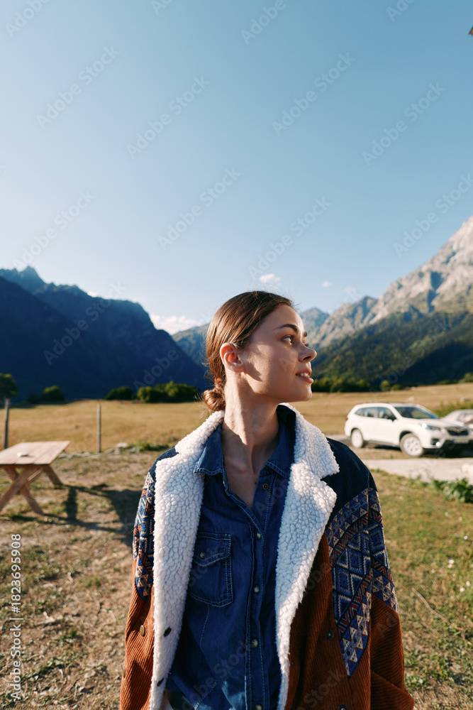 Naklejka premium Woman portrait in mountains near parked car on meadow, wearing warm jacket and denim shirt, travel and nature scene with clear sky, outdoor lifestyle and peaceful scenery