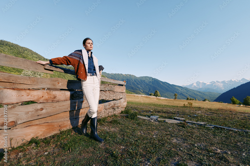 Obraz premium Woman stands by wooden fence in mountain meadow at sunrise, wearing jacket, scarf and rubber boots. Landscape view with rolling hills and clear blue sky for outdoors leisure.
