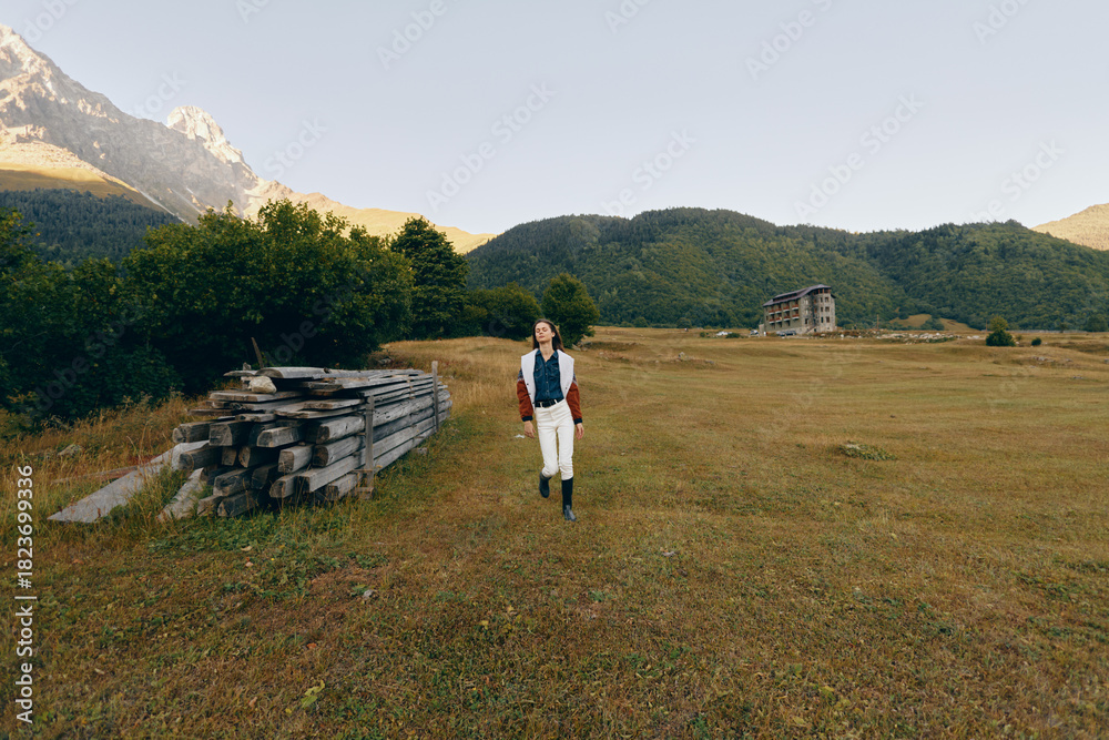 Fototapeta premium Woman walking across a wide meadow and open field with distant mountains in the background, nature landscape in a rural setting, casual outfit with boots and jacket for outdoor exploration.