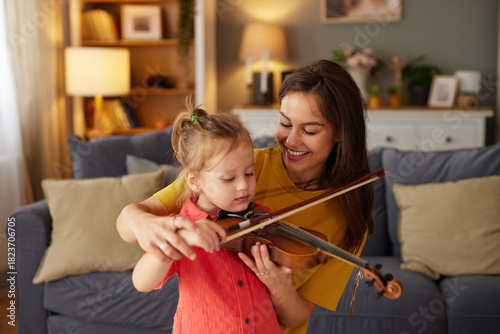A mother smiles while guiding her young daughter in playing the violin. The family is having fun together in their comfortable living room, filled with warmth and love.