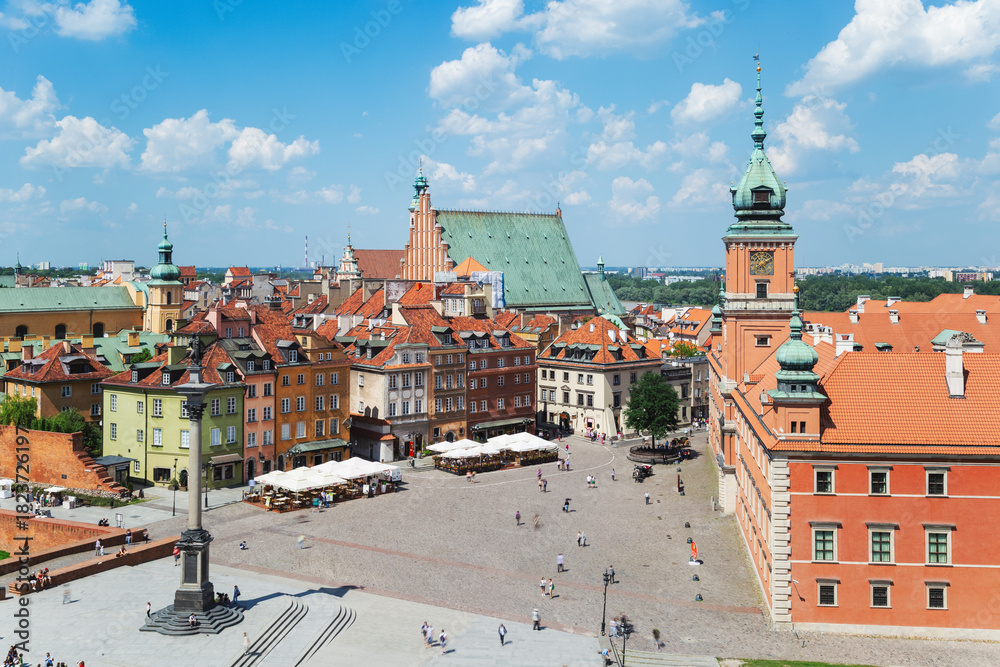 Fototapeta premium A sunny view of Warsaw’s Castle Square with colorful historic buildings, a central column monument, cafés, and people enjoying the lively square under a blue sky.