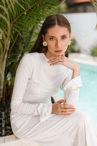 Woman in a long white dress sitting and posing gracefully beside tropical greenery and a pool