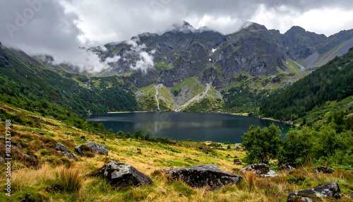 Serene lake mirroring majestic, cloud-veiled mountains, framed by lush greenery and rocky terrain under a gray sky