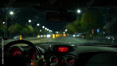 Car interior view of driving through a city streets at night. Unrecognizable male driver, blurry wide angle view of the street and tail lights, rain drops on the windshield, real time video