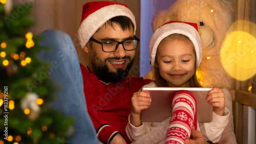 Happy father and little daughter use tablet sitting by Christmas tree. Family leisure time during holidays New Year and Christmas