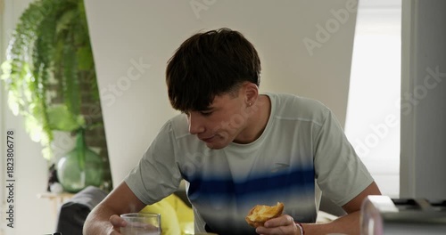 A young Caucasian man is enjoying a moment of tranquillity in his morning routine, drinking coffee with milk from a glass while reading on his phone at the kitchen counter.