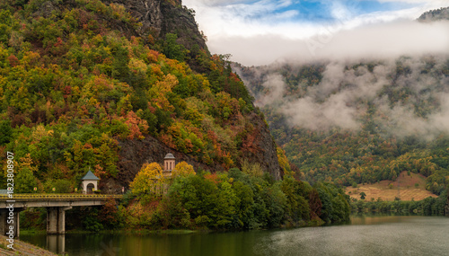Picturesque autumn landscape in the Olt Valley, Romania, with vibrant fall foliage on the mountains. A small historic church and a bridge span the calm Olt River, surrounded by misty, forested hills.