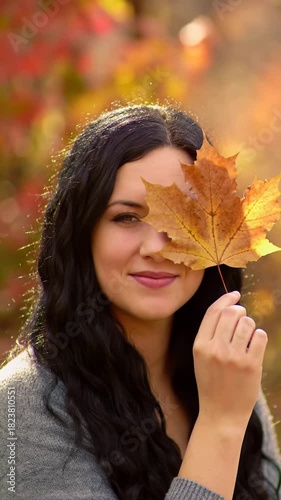 Young caucasian woman with long dark wavy hair, casual top, gently smiles, holding and revealing face with a large dried maple leaf, rich autumn foliage bokeh background. Serene golden hour concept