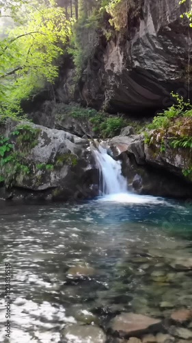 Slow crane video descends, revealing a tranquil waterfall cascading into a clear rocky pool with glistening river stones, embraced by an ancient forest and mossy cliff. Golden hour sunlight creates