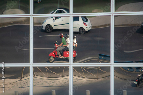 Mirrored windows held by a metal structure reflect a multi-lane urban road on which a white vehicle and a motorcycle with two people who appear to be three pass by