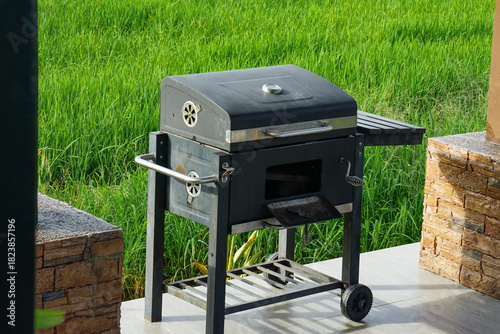 Black metal charcoal grill with lid, air vents, side shelf, and wheeled legs, positioned on a tiled patio between stone pillars overlooking a lush green field.