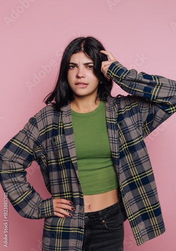 Confused young woman pondering in front of pink background while wearing casual plaid shirt and green top