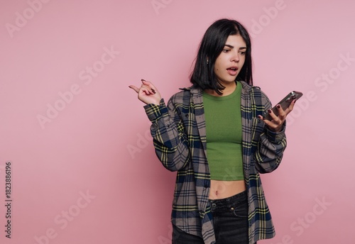 Young woman surprised while texting on smartphone against pink background in casual clothing