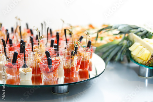 A wide display of assorted appetizer cups arranged on a mirrored platter at a catered event, featuring colorful ingredients and tasting utensils.