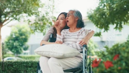 Professional Asian Female Caregiver Hugging Elderly Woman in Wheelchair while relaxing in park. Asian female caregiver take care and talking with Senior woman in wheelchair at park.