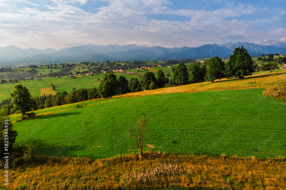 Obraz premium Wide aerial drone panorama of the Tatra Mountains and the village of Sierockie in the Polish Podhale region. Lush green farmlands and rolling hills under a dramatic sky.