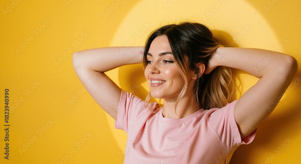 Fototapeta premium Happy young woman smiling with hands in her hair. Cheerful female model in a pink t-shirt posing against a vibrant yellow background with a spotlight. Portrait with copy space for text