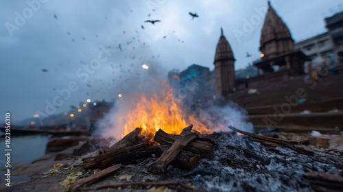 A dramatic scene at Manikarnika Ghat in Varanasi, capturing the ancient cremation rites along the sacred Ganges River. A funeral pyre burns intensely in the foreground, flames risi