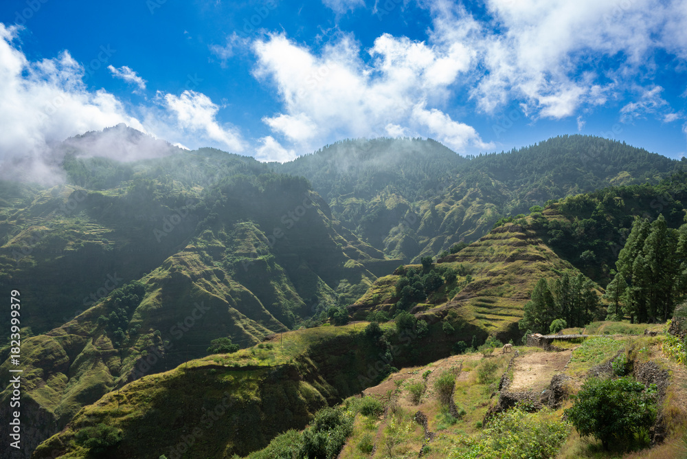 Naklejka premium Scenic terraced mountains of Santo Antão, Cape Verde, with lush green slopes, dramatic valleys and a bright sky, captured in natural sunlight