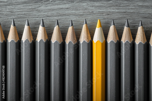 A sharp yellow pencil stands vertically in a row of identical dark gray pencils on a wood-grain surface. Strong symmetry. Individuality, uniqueness, leadership concept.